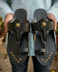 Kolhapur Original in black, a pair held up in an artisan's hands